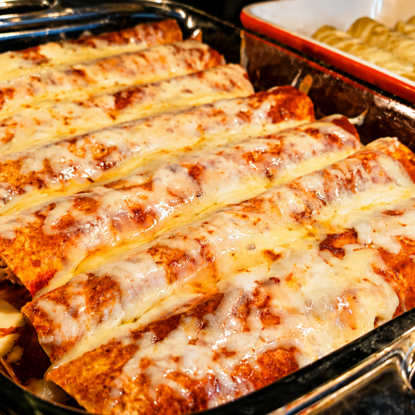 A glass baking dish filled with cheesy chicken enchiladas covered in melted cheese and red sauce sits in an oven next to another dish with a layered baked entrée.