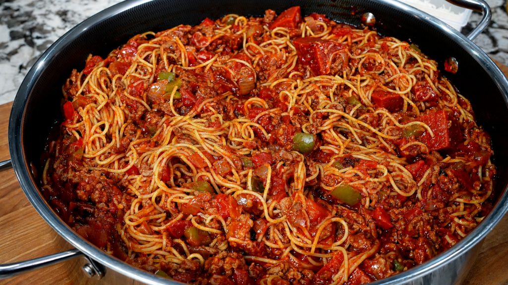 A large pan filled with spaghetti mixed in a rich tomato sauce with ground meat, sliced bell peppers, and visible herbs, set on a wooden surface.