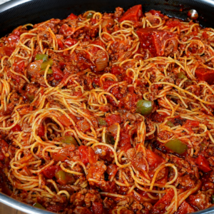 A large pan filled with spaghetti mixed in a rich tomato sauce with ground meat, sliced bell peppers, and visible herbs, set on a wooden surface.