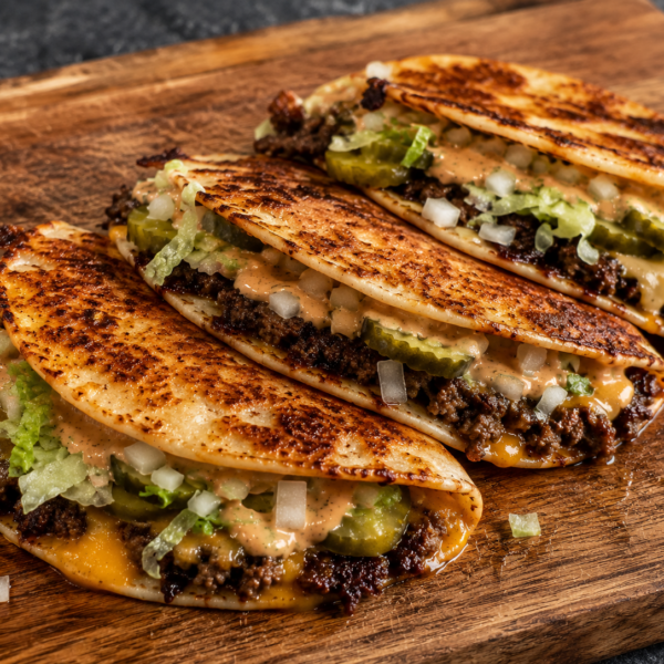 Three cheesy ground beef tacos with lettuce, chopped onions, pickles, and sauce are arranged in a row on a wooden cutting board. The taco shells are golden and slightly crispy.