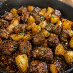 A cast iron skillet filled with browned steak bites and golden roasted potato chunks, seasoned with herbs and spices, sitting on a dark countertop.