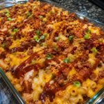 A glass baking dish filled with cheesy macaroni and shredded chicken, topped with barbecue sauce, crispy bacon bits, and chopped green onions on a dark granite counter.