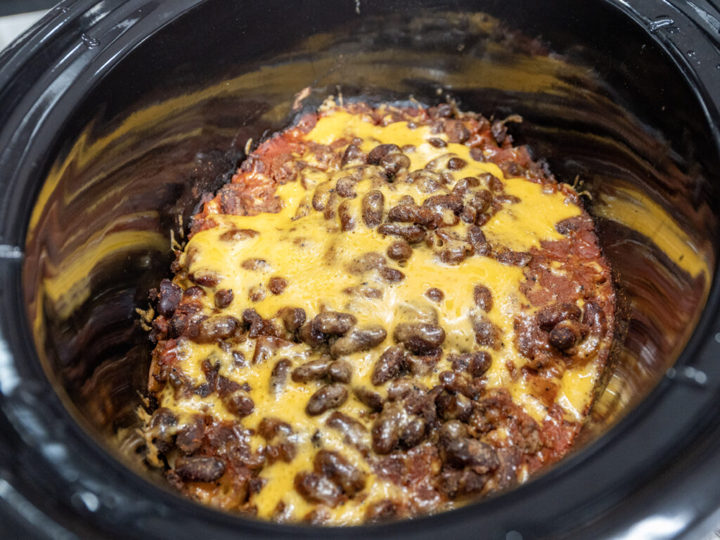 A close-up of chili with beans and melted cheese on top, cooked in a black Crockpot. The chili has a rich, hearty texture with visible tomato sauce, melted yellow cheese, and resembles a Ground Beef Potato Casserole.