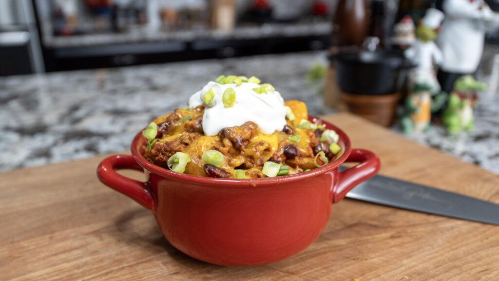 A red bowl filled with chili topped with shredded cheese, a dollop of sour cream, and chopped green onions sits on a wooden cutting board—perfect inspiration for your next Crockpot Ground Beef Potato Cassarole. A knife and blurred kitchen items are in the background.