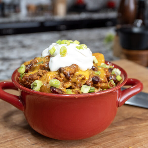A red bowl filled with chili topped with shredded cheese, a dollop of sour cream, and chopped green onions sits on a wooden cutting board&mdash;perfect inspiration for your next Crockpot Ground Beef Potato Cassarole. A knife and blurred kitchen items are in the background.