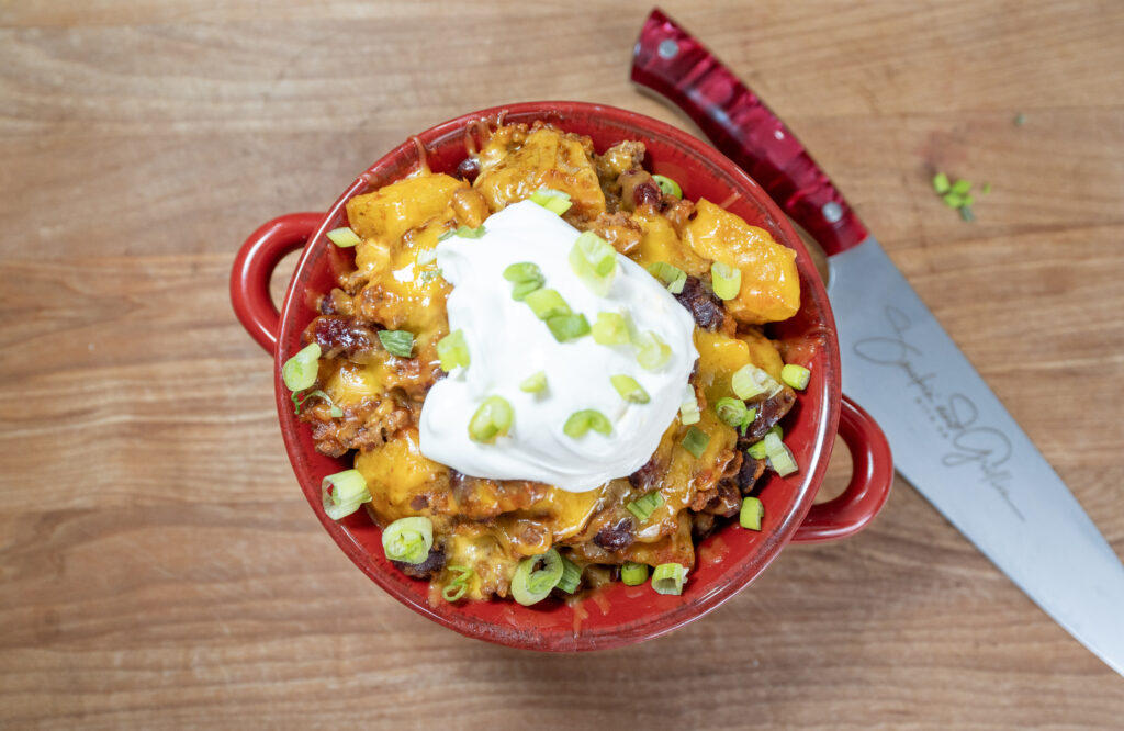 A red bowl filled with a cheesy chili dish reminiscent of Crockpot Ground Beef Potato Casserole is topped with sour cream and chopped green onions, sitting on a wooden surface next to a red-handled kitchen knife.