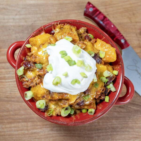 A red bowl filled with a cheesy chili dish reminiscent of Crockpot Ground Beef Potato Casserole is topped with sour cream and chopped green onions, sitting on a wooden surface next to a red-handled kitchen knife.