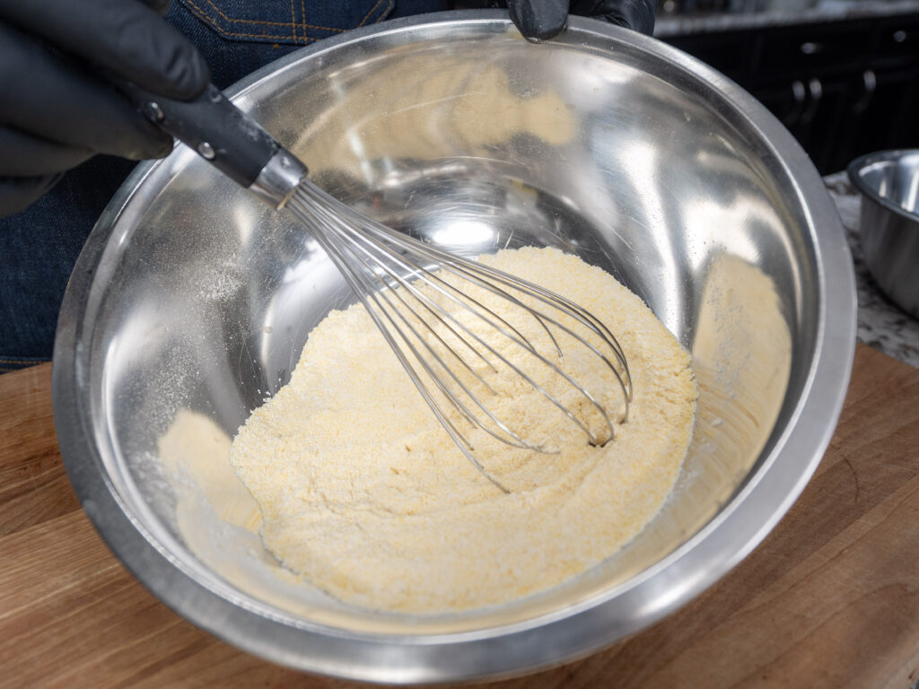 A person wearing black gloves is whisking a yellow dry mixture, likely for cornbread, in a large stainless steel mixing bowl on a wooden surface.