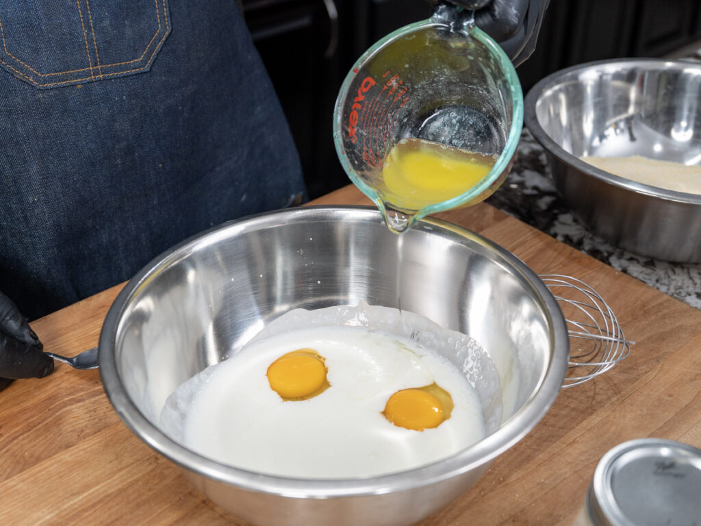A person pours melted butter from a measuring cup into a metal bowl containing yogurt and two raw eggs—ingredients for cornbread—with a whisk and another mixing bowl nearby on a wooden countertop.