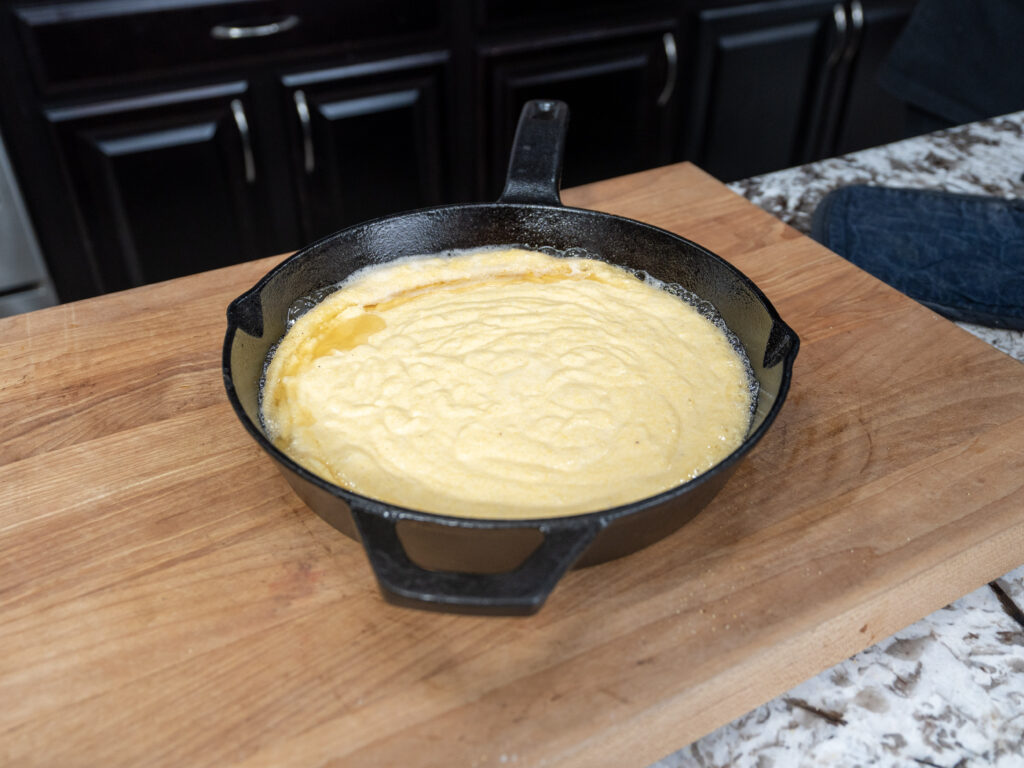 A black cast iron skillet filled with cornbread batter sits on a wooden cutting board in a kitchen with dark cabinets and a granite countertop.