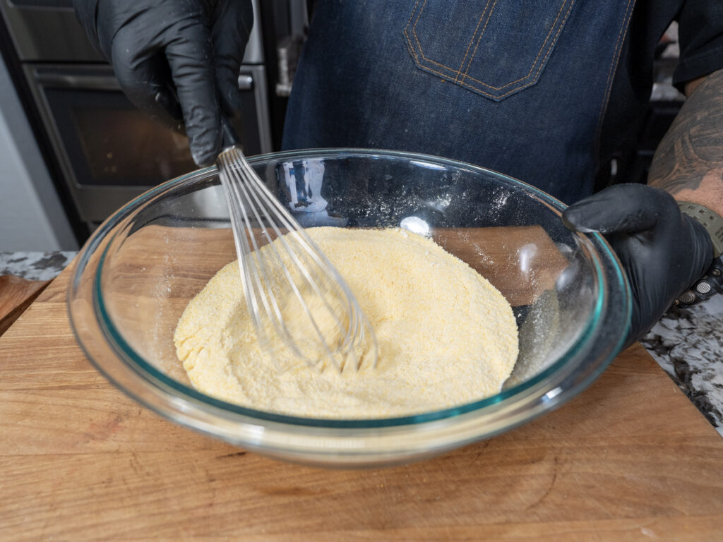 A person wearing black gloves and a denim apron uses a whisk to mix cornmeal for cornbread in a large glass bowl on a wooden surface.