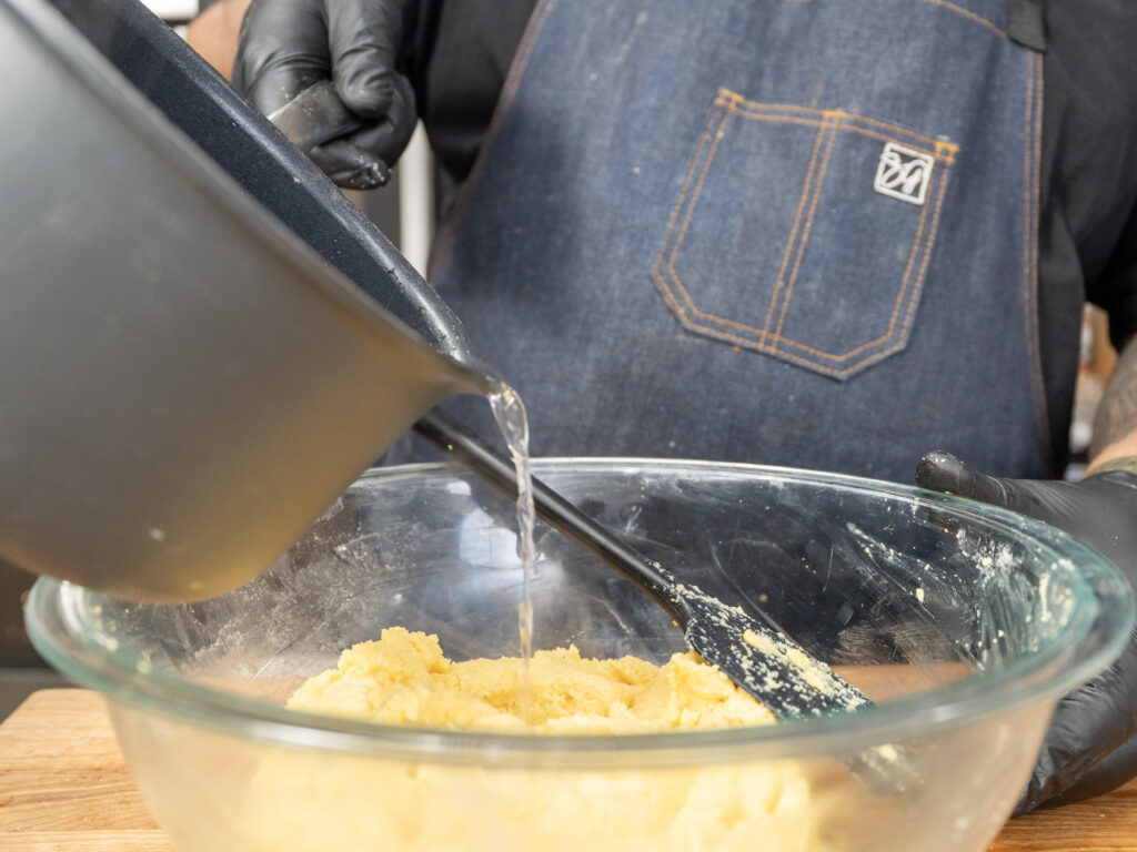 A person wearing a denim apron and black gloves pours water from a black pot into a glass bowl with cornbread dough, using a black spatula to mix the ingredients.