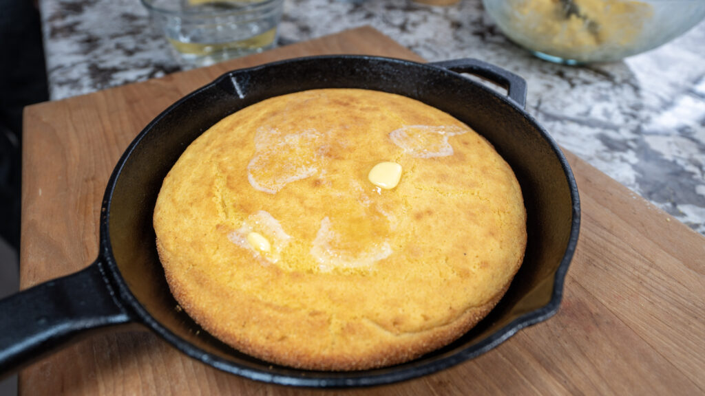 A round, golden cornbread with melted butter on top sits in a black cast iron skillet on a wooden cutting board. A glass of water and mixing bowl are visible in the background, completing this classic cornbread scene.
