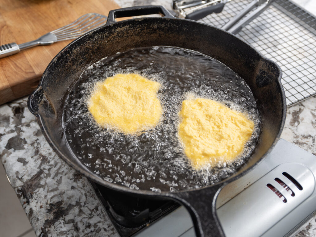 Two pieces of yellow cornbread dough are being deep-fried in hot oil in a black cast iron skillet on a stovetop, with bubbles forming around them. A wooden cutting board and cooling rack are visible nearby.
