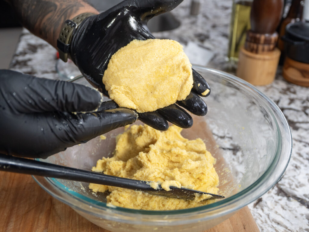 A person wearing black gloves shapes yellow cornbread dough with their hands over a glass bowl filled with more dough, on a kitchen counter with utensils and seasonings in the background.