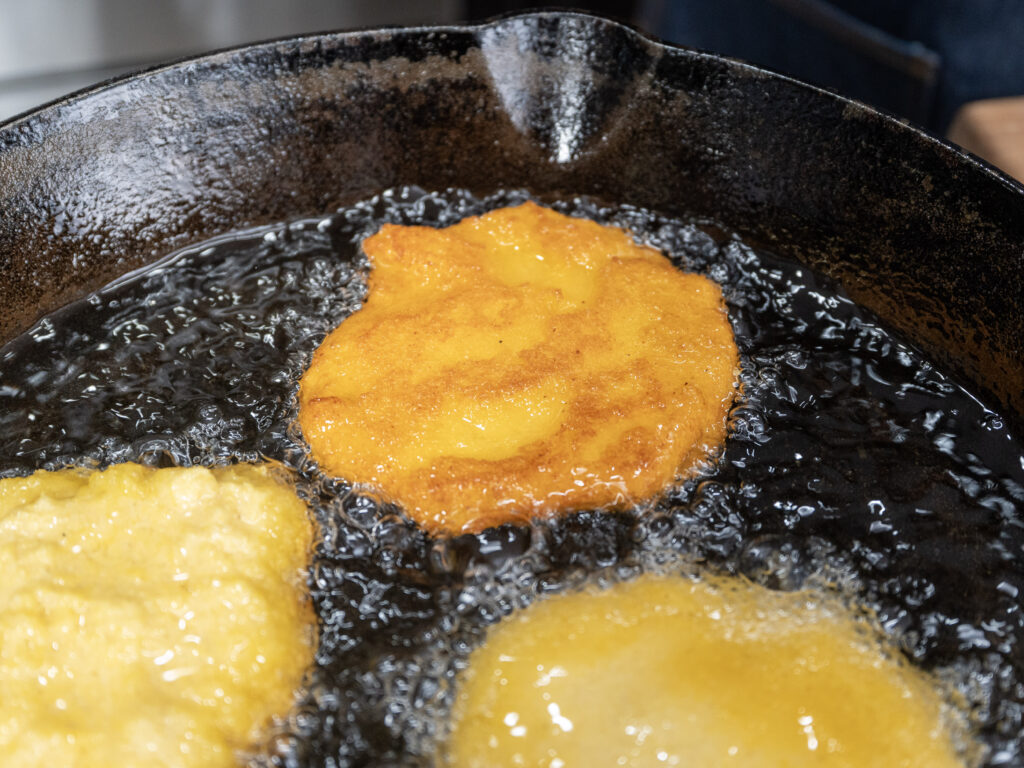 Two pieces of cornbread dough are being fried in hot oil in a black cast iron skillet, with one piece golden brown and the other still light yellow. The oil is bubbling around the dough.