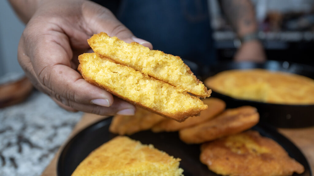 A hand holds two slices of golden, moist cornbread above a plate with more cornbread pieces, while a cast iron skillet sits in the background.