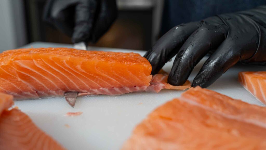 A person wearing black gloves slices fresh salmon fillets on a white cutting board with a sharp knife, preparing them for delicious homemade salmon croquettes.