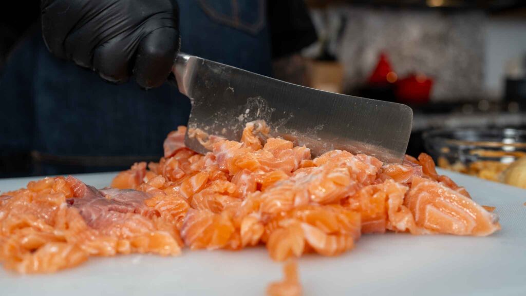 A person wearing a black glove chops raw salmon with a large knife on a white cutting board, preparing the fish in a kitchen setting to make delicious salmon croquettes.