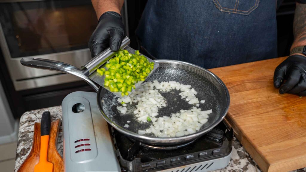 A person wearing black gloves adds chopped green bell pepper to a frying pan with diced onions on a stovetop, preparing ingredients for delicious salmon croquettes. A wooden cutting board and orange spatula are nearby.