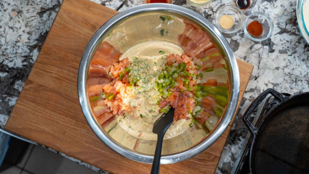A metal mixing bowl on a wooden board holds chopped vegetables, ground meat, herbs, and a creamy sauce for salmon croquettes, being mixed with a black spatula. Various small bowls with spices sit nearby on a marbled countertop.