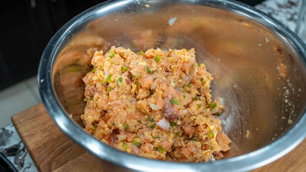 A large metal mixing bowl containing a mixture for salmon croquettes&mdash;chopped raw meat, onions, and green peppers&mdash;sits on a wooden surface in a kitchen.