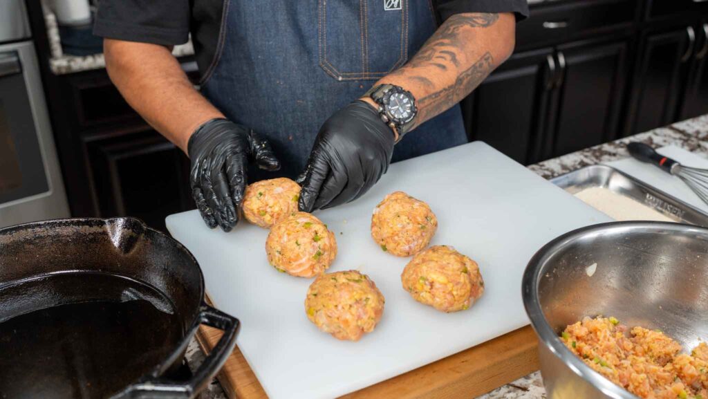 A person wearing black gloves and a dark apron shapes salmon croquettes mixture into round patties on a white cutting board, with more mixture in a bowl and a skillet nearby.