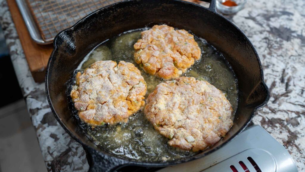 Three salmon croquettes are frying in hot oil in a black cast iron skillet on a stovetop, with bubbling around the edges and a cooling rack visible in the background.