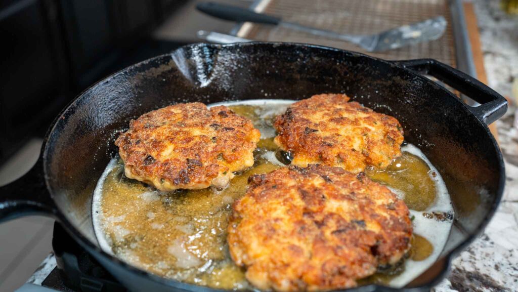 Three golden-brown crab cakes frying in a cast iron skillet with bubbling oil on a stovetop&mdash;reminiscent of classic salmon croquettes. A spatula and cooling rack are visible in the background.