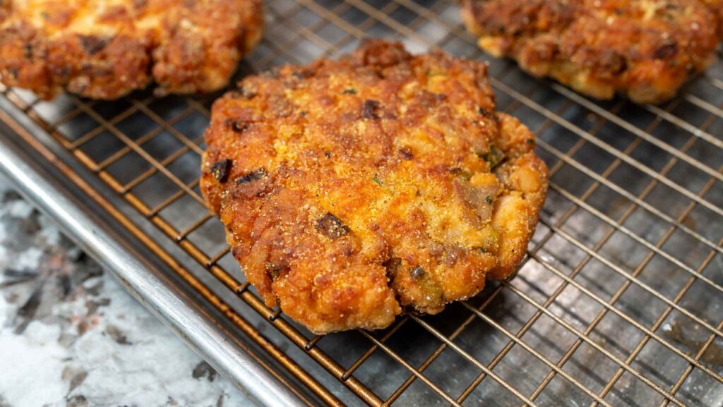 A close-up of a golden-brown, crispy salmon croquette resting on a wire cooling rack over a baking sheet, with other salmon croquettes in the background.