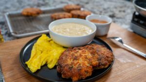 A black plate with yellow scrambled eggs and two crispy salmon croquettes sits beside a bowl of creamy sauce. In the background, more salmon croquettes cool on a wire rack and a cup of brown sauce is nearby on a wooden surface.