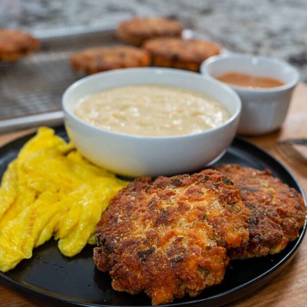 A black plate with yellow scrambled eggs and two crispy salmon croquettes sits beside a bowl of creamy sauce. In the background, more salmon croquettes cool on a wire rack and a cup of brown sauce is nearby on a wooden surface.