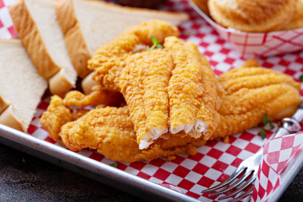 Crispy fried fish fillets served on a red and white checkered paper from the Smokin and Grillin with AB Food Truck, with slices of white bread and a metal fork on the side.