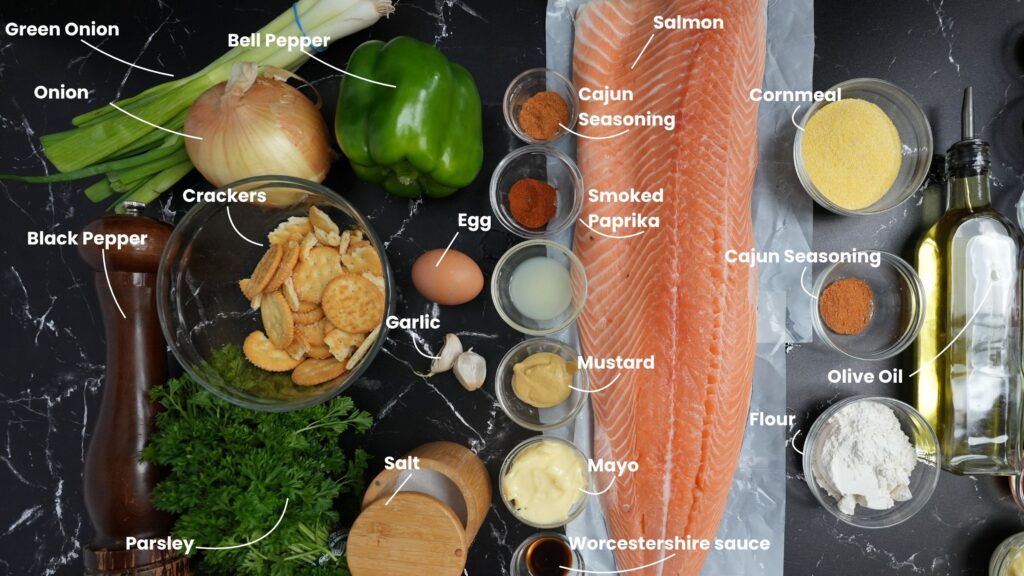 Ingredients for salmon croquettes are laid out on a black surface, including salmon fillet, egg, onion, green onion, bell pepper, parsley, garlic, crackers, seasonings, cornmeal, flour, mayo, mustard, Worcestershire sauce, olive oil and salt.