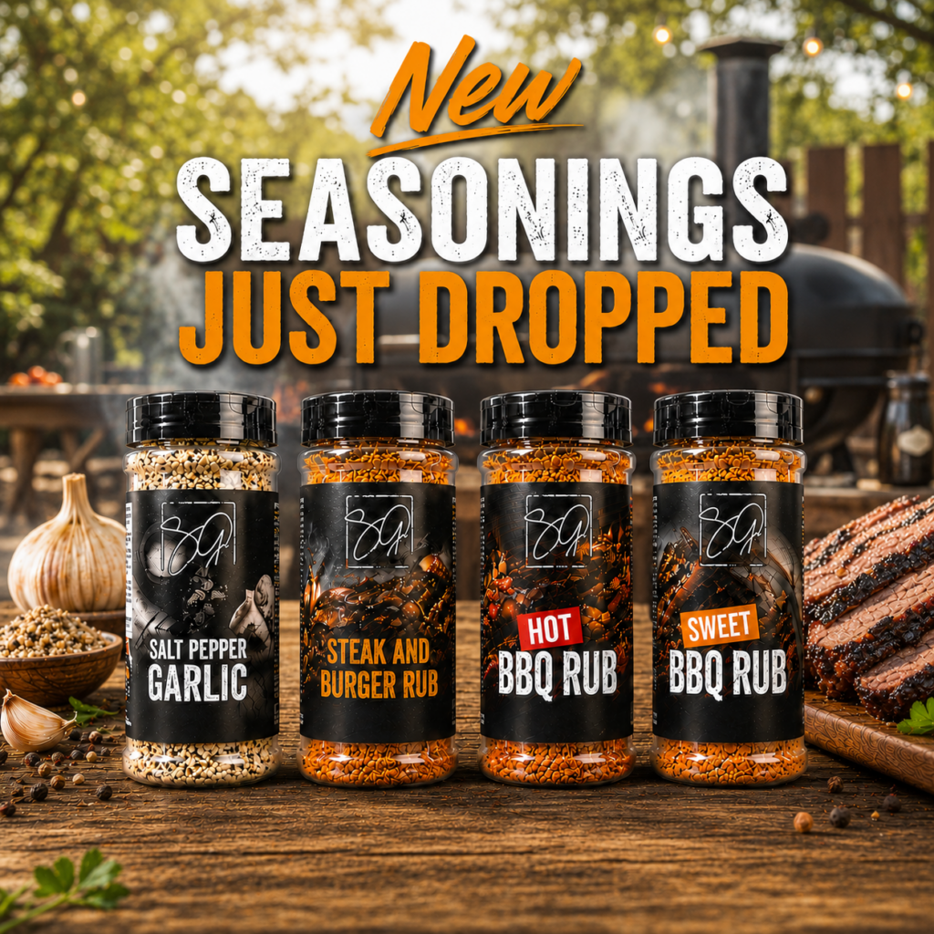 Four bottles of BBQ seasonings—Salt Pepper Garlic, Steak and Burger Rub, Hot BBQ Rub, and Sweet BBQ Rub—are displayed on a wooden table outdoors with a grill, steak, and fresh ingredients in the background. Text reads: New Seasonings Just Dropped.