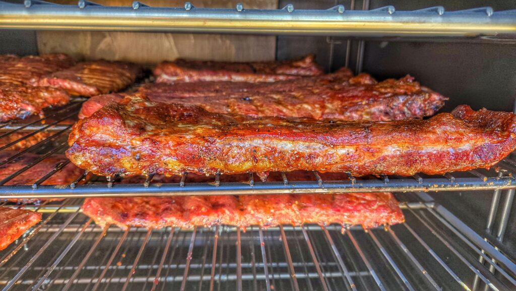 Close-up of racks of seasoned pork ribs cooking on metal grill shelves inside the Smokin and Grillin with AB Food Truck smoker, with a golden-brown, crispy exterior visible on the ribs.