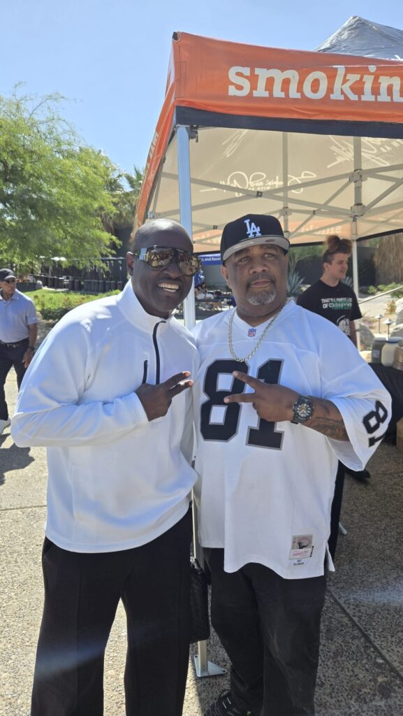 Two men pose and smile outdoors at a food stand. The man on the left wears sunglasses and a white pullover, while the man on the right wears a white Raiders jersey and LA cap. An orange tent and other people are in the background.