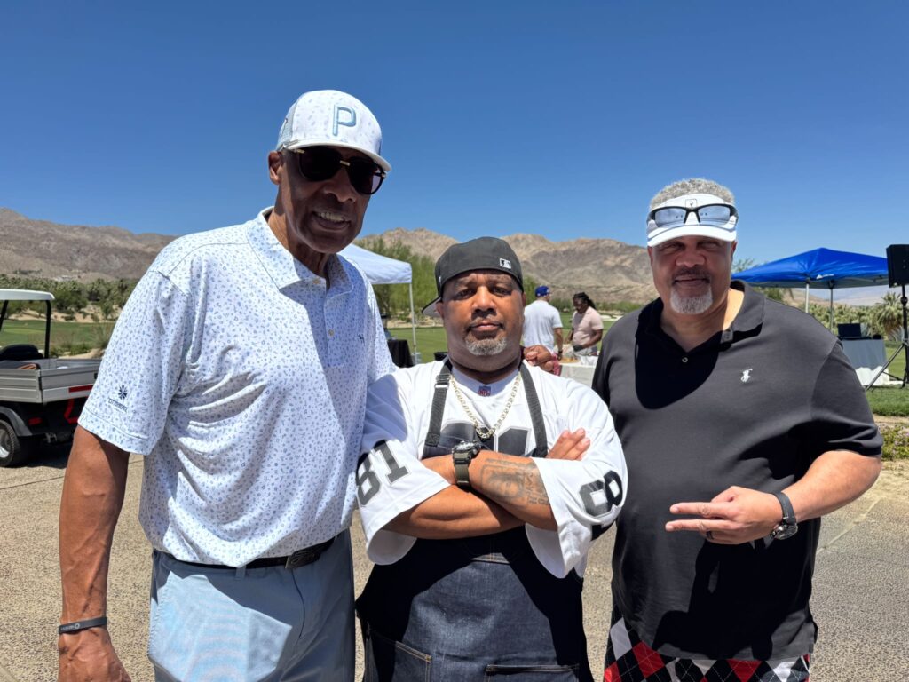 Three men stand outdoors under a clear blue sky with mountains in the background. Two are wearing hats and sunglasses, while the person in the center wears an apron and headscarf. Tents and a vehicle are visible behind them.