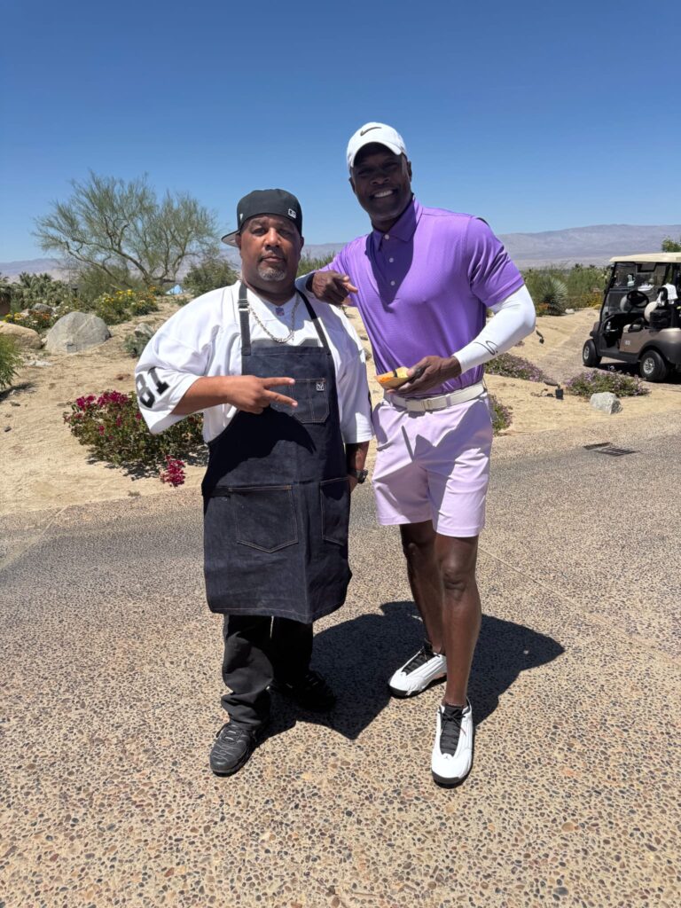 Two men stand outside on a sunny day; one wears a black apron and hat, making a peace sign, while the other is dressed in a purple golf shirt and shorts, holding a golf club. Desert plants and a golf cart are in the background.