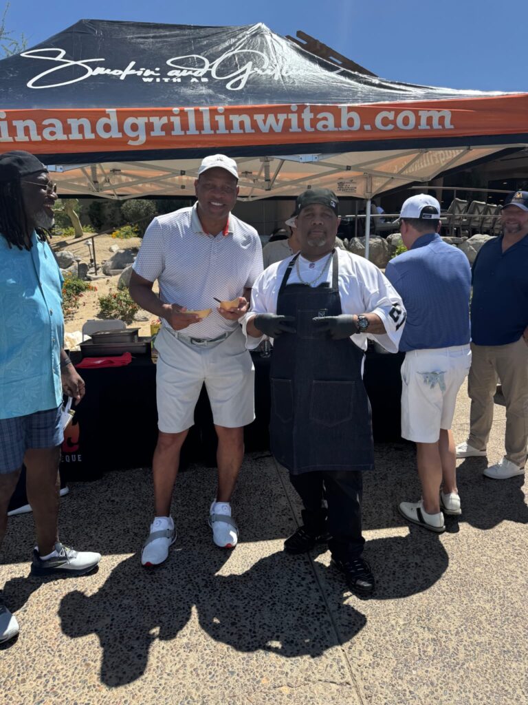 A group of men pose outdoors in front of a grill and BBQ tent, with one man dressed as a chef holding grilling utensils. The others are casually dressed and smiling under a sunny, blue sky.