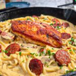 A skillet filled with creamy chicken sausage alfredo pasta, topped with a seared chicken breast, mushrooms, and garnished with chopped parsley.