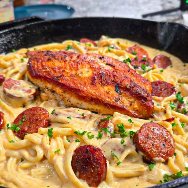 A skillet filled with creamy chicken sausage alfredo pasta, topped with a seared chicken breast, mushrooms, and garnished with chopped parsley.