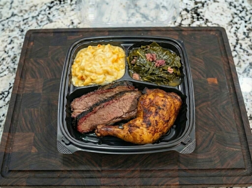 A takeout container from AB&rsquo;s BBQ Fusion food trailer with baked chicken leg, sliced brisket, macaroni and cheese, and collard greens sits on a dark cutting board on a speckled countertop.