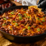 A skillet filled with cheesy bbq baked bean dip topped with ground meat, bacon, and sliced green onions. A gloved hand lifts a tortilla chip dripping with melted cheese from the dip, while more chips are scattered nearby.