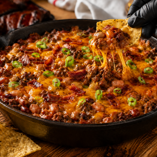 A skillet filled with cheesy bbq baked bean dip topped with ground meat, bacon, and sliced green onions. A gloved hand lifts a tortilla chip dripping with melted cheese from the dip, while more chips are scattered nearby.