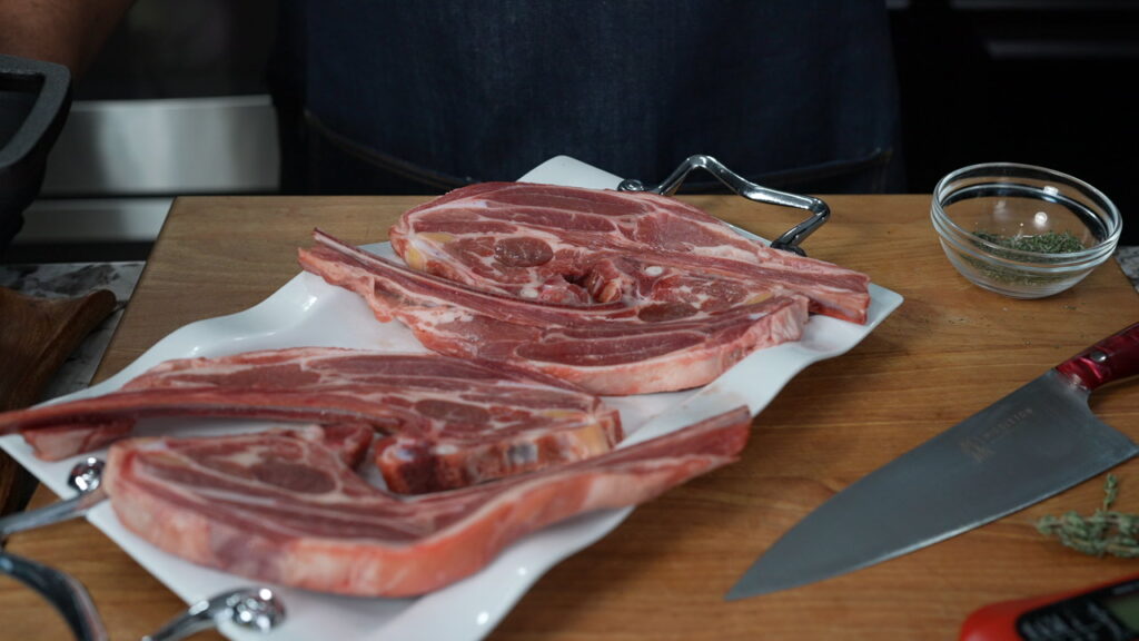 Two raw tomahawk steaks are placed on a white platter atop a wooden cutting board. A chef’s knife, a small bowl of herbs for garlic butter lamb chops, and part of a person in a dark apron are visible in the background.