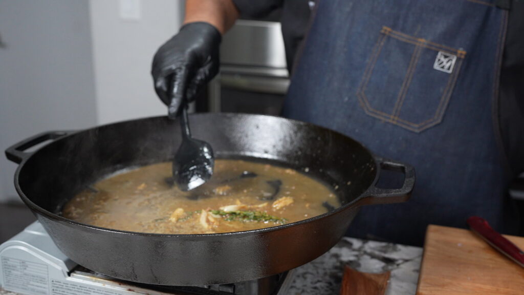 A person wearing a blue apron and black glove stirs garlic butter lamb chops in a large black cast iron skillet filled with broth and herbs on a stovetop. A wooden cutting board is visible on the right.