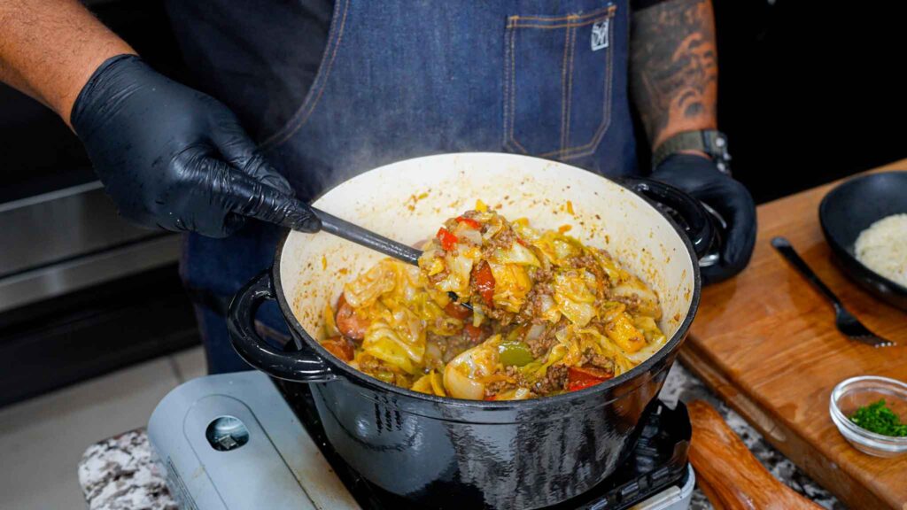 A person wearing black gloves and a blue apron stirs a hearty stew inspired by a dirty cabbage recipe, with vegetables and meat in a black pot on the stovetop, plus a bowl of rice and seasonings nearby on a wooden board.