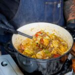 A person wearing black gloves and a blue apron stirs a hearty stew inspired by a dirty cabbage recipe, with vegetables and meat in a black pot on the stovetop, plus a bowl of rice and seasonings nearby on a wooden board.