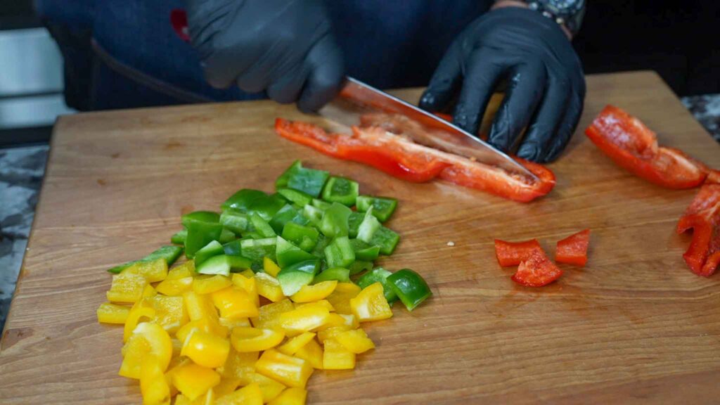A person wearing black gloves is slicing a red bell pepper on a wooden cutting board, with chopped yellow and green bell peppers next to it—perfect ingredients for a vibrant dirty cabbage recipe.